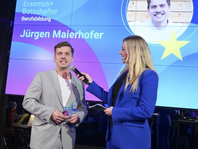 Mann in grauem Anzug spricht in Mikro, das blonde Frau mit blauem Anzug hält