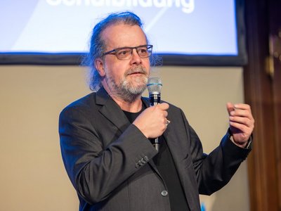 Mann mit grauem Bart und Brille spricht mit Mikrofon in der Hand