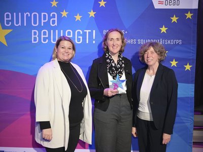 Drei Frauen stehen vor einem Transparent mit der Aufschrift "Europa bewegt" und lachen in die Kamera.