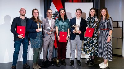 Gruppenfoto der Preisträgerinnen und Preisträger: Sieben Personen stehend teilweise mit roten Urkundenmappen in der Hand