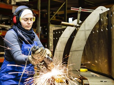 Junge Frau mit Kopftuch, Ohrenschützer und Schutzbrille schneidet Metall, die Funken springen