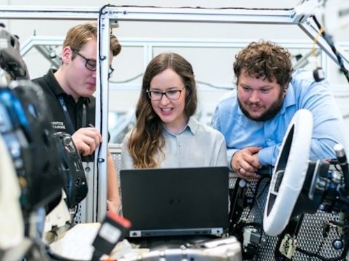 2 junge Männer neben einer jungen Frau mit Laptop - alle betrachten den Innenraum einer offenen Autokarrosserie im Rohbau