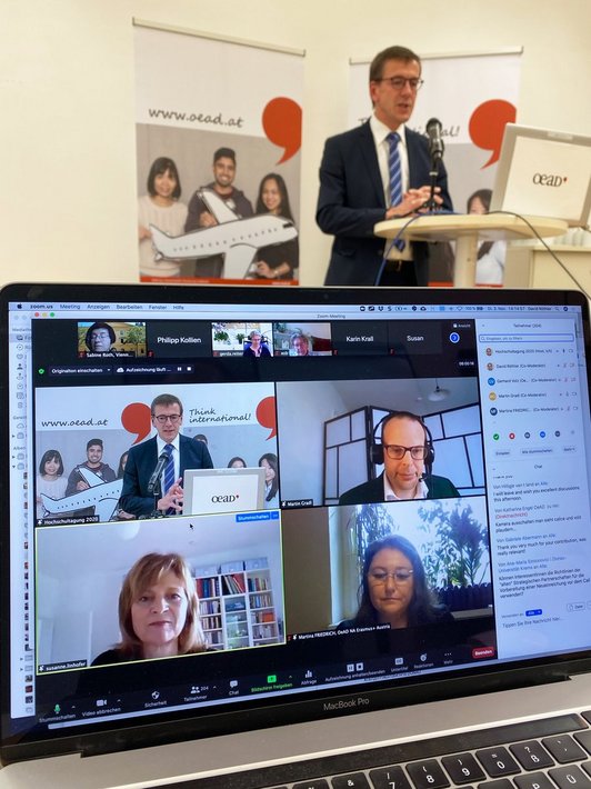 A lecturer stands at a standing desk and below in the foreground is a screen showing images of four people