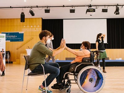 Ein Mann sitzt auf einem Stuhl. Ihm gegenüber sitzt eine Frau im Rollstuhl. Beide heben die Hände und schlagen freundschaftlich mit einander ein. Vielleicht begrüßen sie sich. Die Szene spielt in einem Seminarraum. Im Hintergrund ist eine Bühne und eine Leinwand zusehen, sowie ein paar andere SeminarteilnehmerInnen.