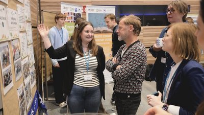Frau steht vor Pinnwand und zeigt auf die Pinnwand, neben ihr stehen vier Personen und hören ihr zu