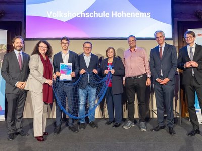 Männer und Frauen in festlicher Kleidung lachend. Eine Person hält Urkunde, eine Person einen Stern, drei Personen halten ein blaues Fischernetz 