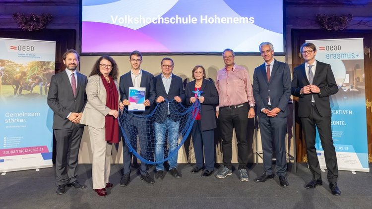 Männer und Frauen in festlicher Kleidung lachend. Eine Person hält Urkunde, eine Person einen Stern, drei Personen halten ein blaues Fischernetz 