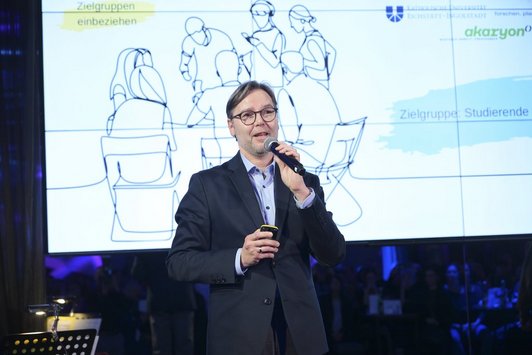 Ein Mann steht auf der Bühne, in der Hand hält er ein Mikrofon, hinter ihm ein Screen
