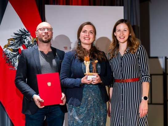 Drei Personen in festlicher Kleidung vor Österreichfahne: Mann mit roter Mappe, Frau mit Trophäe aus Holz und Ministerin Bauer 