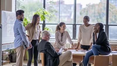 Gruppe von Personen in angeregter Unterhaltung in einem Workshop-Raum mit hohen Glasfronten. Männer und Frauen unterschiedlichen Alters und Hautfarbe, manche sitzend, manche stehend