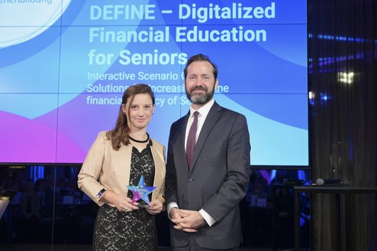 Eine Frau und ein Mann stehen auf der Bühne, hinter ihnen ein Screen mit dem Schriftzug „DEFINE – Digitalized Financial Education for Seniors“.