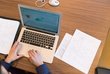 Aufgeklappter Laptop auf Tisch mit Händen an Tastatur, daneben weiße Notizblätter