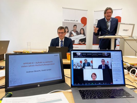 One speaker stands at a standing desk and speaks, another sits at the table with a laptop in front of him and two screens in the foreground, one with the title of the current lecture and the name of the speaker and on the other with pictures of three people