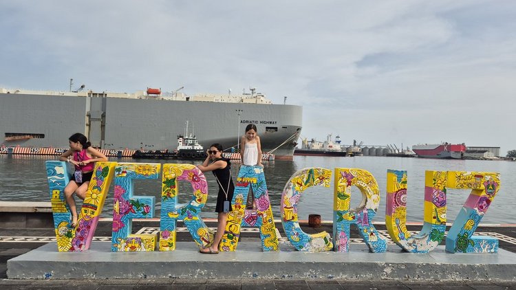 Drei Mädchen posieren an einem bunten, künstlerisch gestalteten Schriftzug 'VERACRUZ' am Hafen. Im Hintergrund liegt ein großes Frachtschiff mit dem Namen 'ADRIATIC HIGHWAY' im Wasser. Der Himmel ist leicht bewölkt, und weitere Schiffe sind in der Ferne zu sehen. Der Schriftzug ist farbenfroh mit folkloristischen Motiven bemalt.