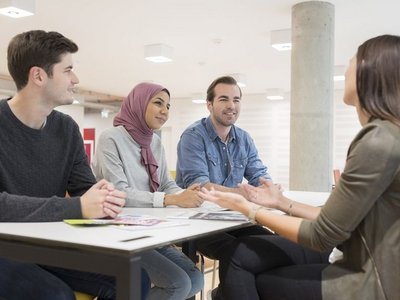 Bild von Studierenden der Wirtschaftsuniversität Wien.  Zwei Männer und zwei Frauen sitzen um einen eckigen Tisch und unterhalten sich. Eine Frau trägt ein fliederfarbiges Kopftuch, alle Studierenden lächeln.