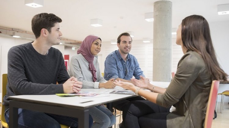 Bild von Studierenden der Wirtschaftsuniversität Wien.  Zwei Männer und zwei Frauen sitzen um einen eckigen Tisch und unterhalten sich. Eine Frau trägt ein fliederfarbiges Kopftuch, alle Studierenden lächeln.