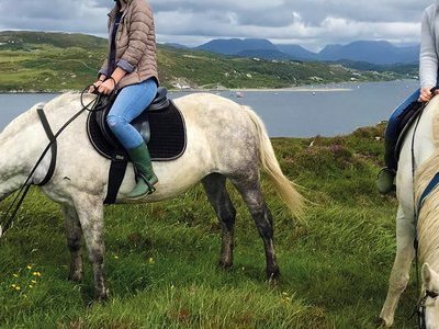 Zwei junge Frauen reiten auf zwei weißen Pferden über eine grüne Wiese, im Hintergrund ist ein See
