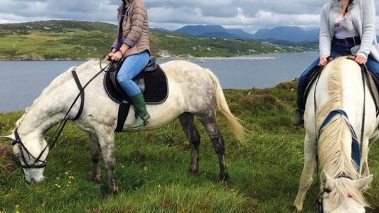 Zwei junge Frauen reiten auf zwei weißen Pferden über eine grüne Wiese, im Hintergrund ist ein See
