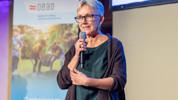 Botschafterin Veronika Drexel vor Roll-Up sprechend mit einem Mikrophon in der Hand