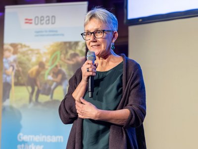 Botschafterin Veronika Drexel vor Roll-Up sprechend mit einem Mikrophon in der Hand