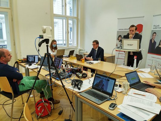 Tisch mit drei Vortragenden, zwei davor sitzen vor jeweils einem Laptop, einer steht am Pult, und ein Techniker, der vor einem Schaltpult und mehreren Bildschirmen sitzt