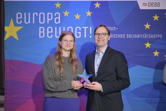 Ein Mann und eine Frau stehen mit einem Pokal in Sternform vor einem Transparent mit der Aufschrift „Europa bewegt“