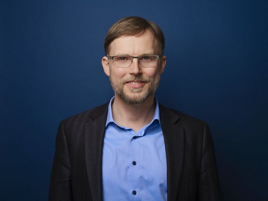 Porträtbild: Dunkelblonder Mann mit Brille lacht frontal in die Kamera. Er trägt ein hellblaues Hemd und ein schwarzes Sakko, der Hintergrund ist dunkelblau. 