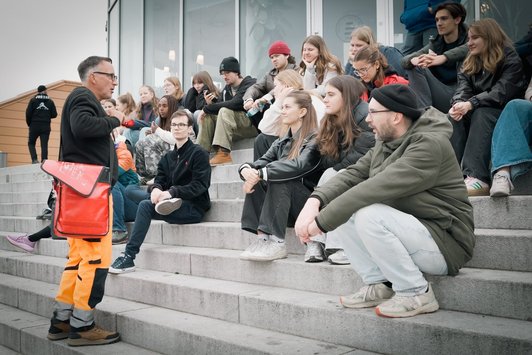 Gruppe junger Menschen sitzt auf Stiege und hört altem Mann zu