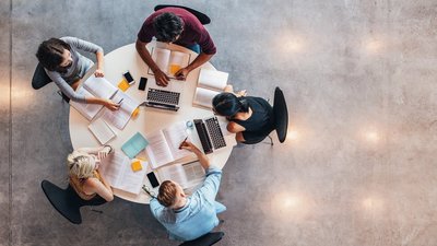 Fünf junge Studierende an einem Runden Tisch, vor ihnen liegen Bücher, Laptops, von oben fotografiert