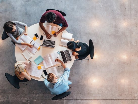 Fünf junge Studierende an einem Runden Tisch, vor ihnen liegen Bücher, Laptops, von oben fotografiert