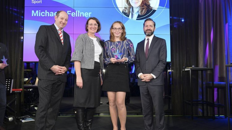 Vier Personen stehen auf der Bühne, eine Person hält einen Pokal. Hinten auf der Leinwand steht „Erasmus+ Botschafterin Sport Michaela Fellner“