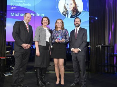 Vier Personen stehen auf der Bühne, eine Person hält einen Pokal. Hinten auf der Leinwand steht „Erasmus+ Botschafterin Sport Michaela Fellner“