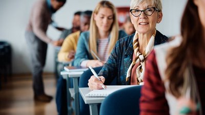 Eine Reihe sitzender Personen im Klassenzimmer. Im Fokus ältere lächelnde Frau mit kurzen Haaren und Brille 