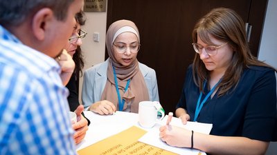 Personen stehen um Tisch herum zusammen, eine Person macht Notizen, die anderen beobachten aufmerskam