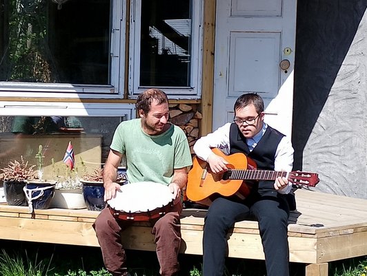 Zwei Männer sitzen auf Holzveranda im Freien. Einer spielt Gitarre, der andere hält eine Trommel in der Hand