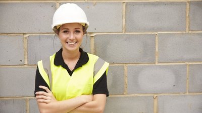 Junge Frau mit weißem Helm und gelber Warnweste steht für einer Ziegelmauer und lächelt in die Kamera