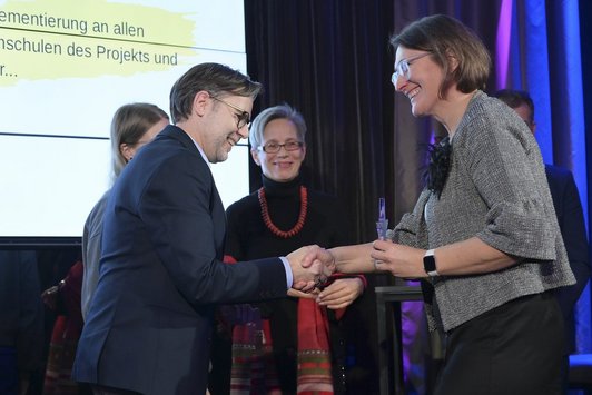 Ein Mann und eine Frau schütteln sich die Hände. Die Frau überreicht dem Mann einen Pokal in Sternform