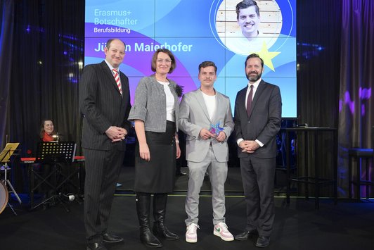 Drei Männer und eine Frau stehen auf der Bühne, Mann hält Auszeichnung 