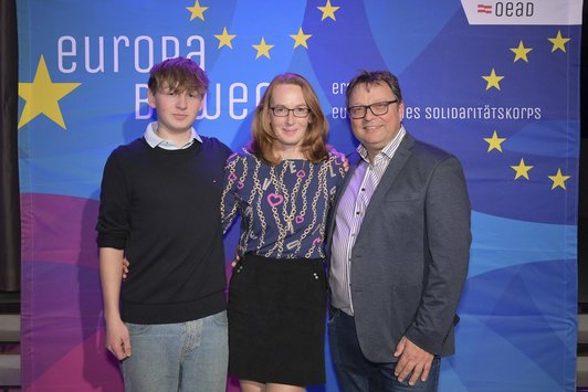 Drei Personen stehen vor einem Transparent mit der Aufschrift "Europa bewegt" und lächeln in die Kamera