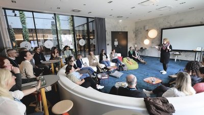 Großer Raum gefüllt mit Personen, eine Dame spricht und steht in der Mitte