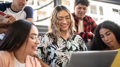 Gruppe von Studierenden sitzt gemeinsam auf Stufen und blicken auf den Laptop einer Kollegin