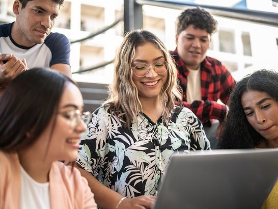 Gruppe von Studierenden sitzt gemeinsam auf Stufen und blicken auf den Laptop einer Kollegin