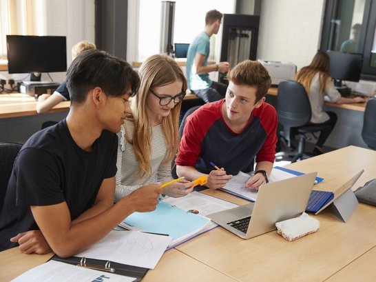 Drei junge Personen gemeinsam in eine Arbeit vertieft sitzen an Tisch und vor ihnen Hefte und Laptops