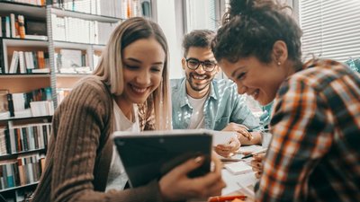 Drei junge Studierende in Freizeitkleidung sitzen an einem Tisch in Bibliothek und blicken lachend auf ein Tablet