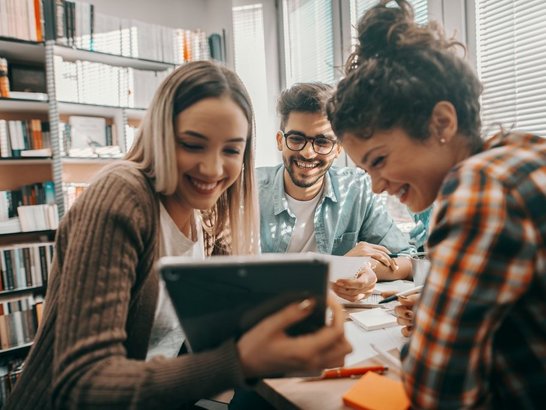 Drei junge Studierende in Freizeitkleidung sitzen an einem Tisch in Bibliothek und blicken lachend auf ein Tablet