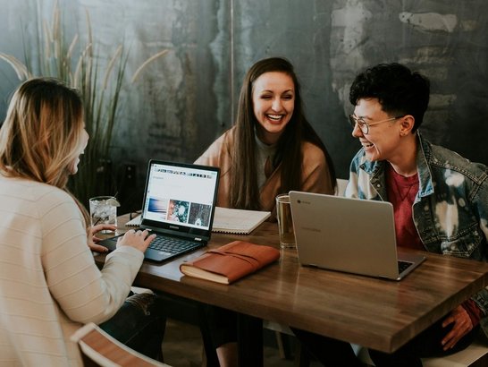 Drei Personen sitzen an einem Holztisch in einem modernen Café und arbeiten gemeinsam an Laptops. Sie lachen und wirken entspannt. Auf dem Tisch liegen Notizbücher, ein Glas Wasser und ein weiteres Getränk. Im Hintergrund sind Pflanzen und eine graue Wand zu sehen.