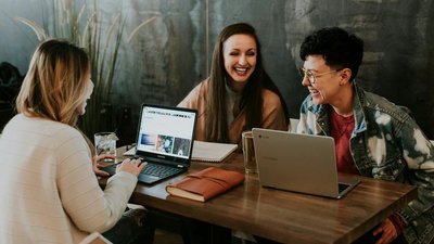 Drei Personen sitzen an einem Holztisch in einem modernen Café und arbeiten gemeinsam an Laptops. Sie lachen und wirken entspannt. Auf dem Tisch liegen Notizbücher, ein Glas Wasser und ein weiteres Getränk. Im Hintergrund sind Pflanzen und eine graue Wand zu sehen.