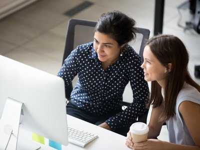 Multiracial colleagues indian and caucasian young women having coffee break sitting together at desk in office. Diverse students interns friends looks at pc screen talking discussing working moments