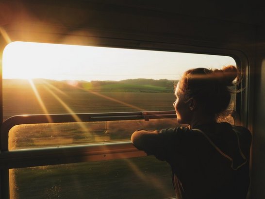 Junge Person steht an offenen Zugfenster und blickt in den Sonnenaufgang 