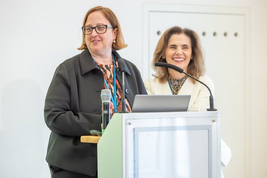Alexandra Enzi am Redepult, Carin Dániel Ramírez-Schiller steht schräg hinter ihr lächelnd.